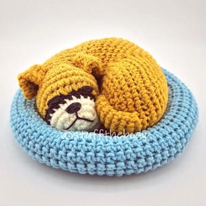 Può includere: Un giocattolo per cani all'uncinetto in posizione rannicchiata, appoggiato su un letto azzurro all'uncinetto. Il cane è giallo dorato con un muso bianco e nero. Il letto è di forma circolare.