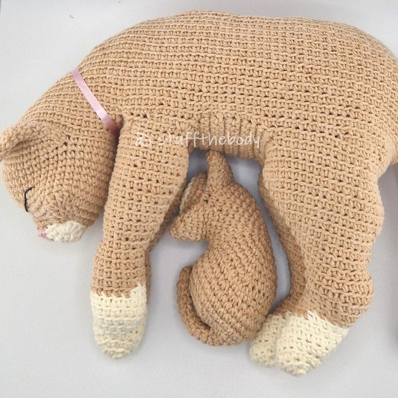 May include: A crocheted cat-shaped pillow in tan with a smaller kitten-shaped pillow nestled beneath it. The larger cat has white paws and a pink ribbon tied around its neck. The kitten is also tan.