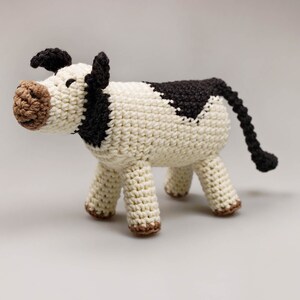 May include: A crocheted stuffed cow toy with a white body, black spots, and brown face. The cow has a long tail and four legs.