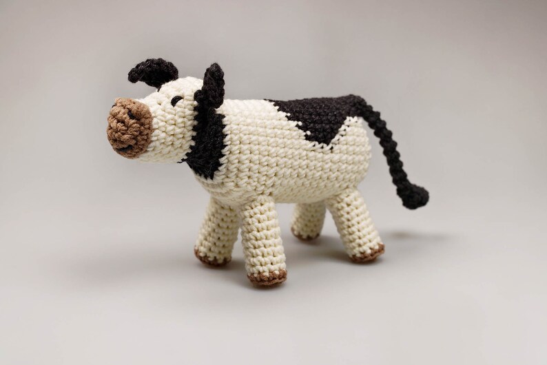 May include: A crocheted stuffed cow toy with a white body, black spots, and brown face. The cow has a long tail and four legs.