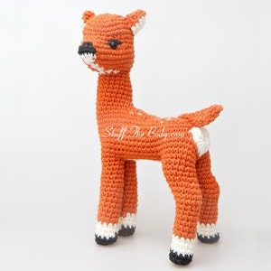 May include: A crocheted deer toy with white spots and black eyes. The deer is standing on a white surface.