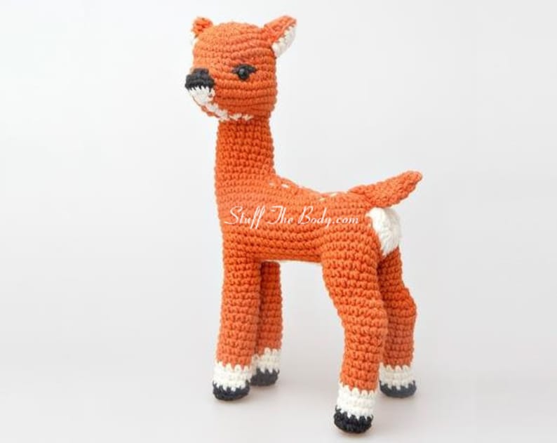 May include: A crocheted deer toy with white spots and black eyes. The deer is standing on a white surface.