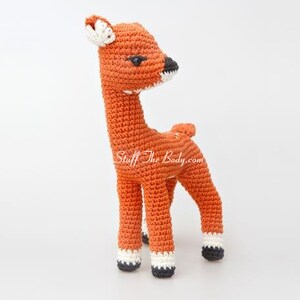 May include: A crocheted deer toy with orange fur, black eyes, and white accents on the hooves. The deer is standing on a white surface.