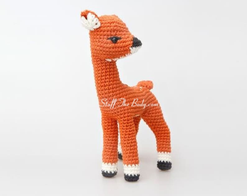 May include: A crocheted deer toy with orange fur, black eyes, and white accents on the hooves. The deer is standing on a white surface.