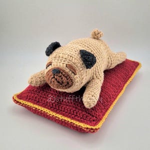 Op de afbeelding: Een gehaakt mopshond-speeltje in beige, met zwarte oren en een bruine snuit, rustend op een rood kussen met gele rand. De handgemaakte hond ligt ontspannen. Het speelgoed is een zacht, knuffelbaar item.