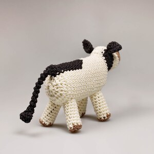 May include: A crocheted stuffed cow toy with a white body, black spots, and a long black tail. The cow is standing on a light gray surface.