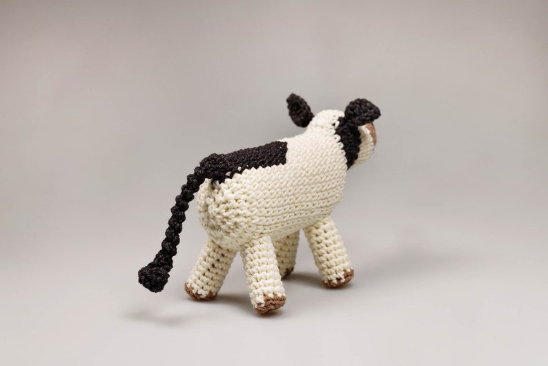 May include: A crocheted stuffed cow toy with a white body, black spots, and a long black tail. The cow is standing on a light gray surface.