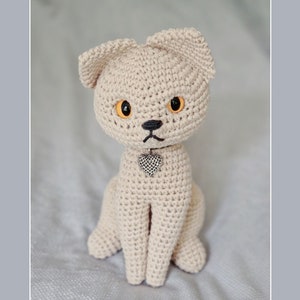 Puede incluir: Un juguete de gato de ganchillo beige con ojos naranjas y un dije de corazón plateado. El gato está sentado sobre una superficie gris claro.