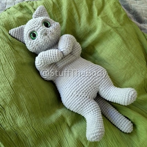 May include: A handmade, light gray crochet cat doll with bright green eyes. The cat is lying on its back on a green fabric, with its paws crossed and tail extended. The doll has a relaxed pose.
