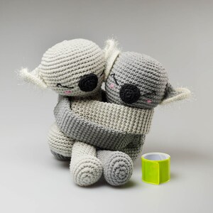 May include: Two gray crocheted koala plushies hugging each other. The koala on the left is white and the koala on the right is gray.