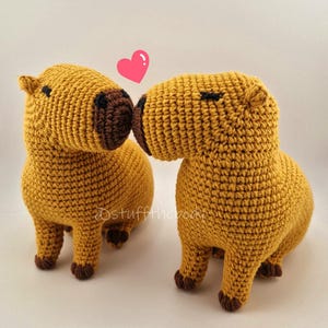 Puede incluir: Dos capibaras de ganchillo hechos a mano en una pose de beso, con un corazón rosa encima. Los capibaras son de color amarillo dorado con detalles marrones, incluyendo sus narices, pies y ojos. La imagen tiene una estética suave y cálida.