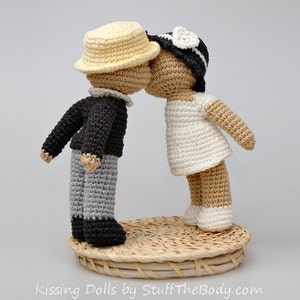 Peut inclure: Deux poup&eacute;es en crochet s'embrassant. La poup&eacute;e de gauche porte un pull noir, un pantalon gris et un chapeau jaune. La poup&eacute;e de droite porte une robe blanche et un chapeau noir et blanc. Les poup&eacute;es sont debout sur une base tiss&eacute;e.