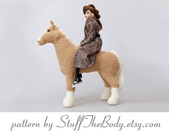 May include: A crocheted horse with a white mane and tail, being ridden by a doll wearing a leopard print coat. The doll has brown hair and is wearing black shoes. The horse is tan and white.