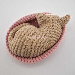 May include: A handmade crochet cat toy in a pink bed. The cat is light brown, curled up, and has pointed ears. The bed is a light pink, round shape with a textured surface. The toy is on a white surface.