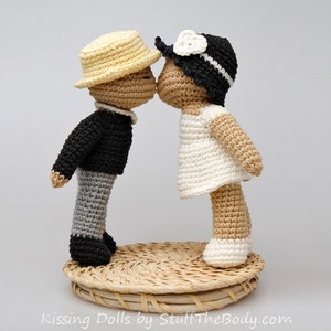 Peut inclure: Deux poup&eacute;es en crochet, l'une portant un chapeau jaune et une veste noire, l'autre portant une robe blanche et un chapeau noir, s'embrassent. Les poup&eacute;es sont debout sur un panier tiss&eacute;.