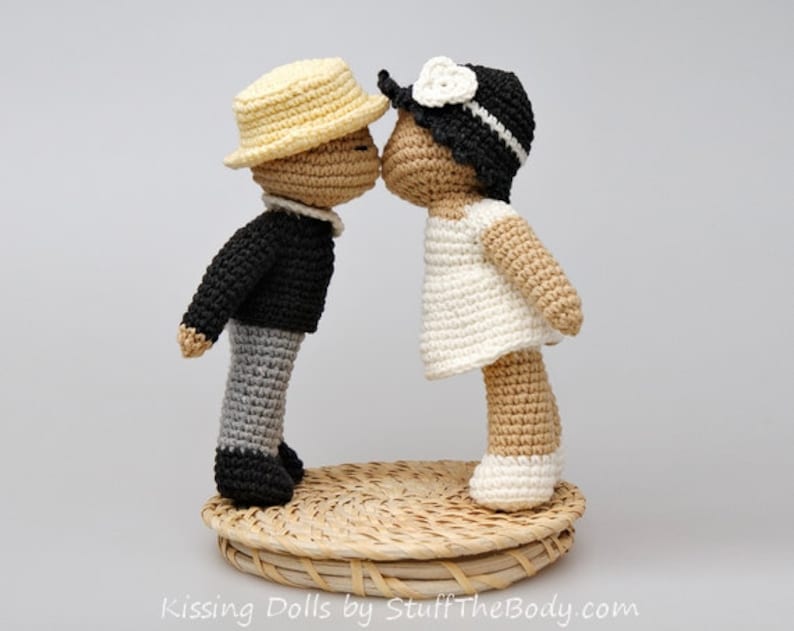 Peut inclure: Deux poup&eacute;es en crochet, l'une portant un chapeau jaune et une veste noire, l'autre portant une robe blanche et un chapeau noir, s'embrassent. Les poup&eacute;es sont debout sur un panier tiss&eacute;.