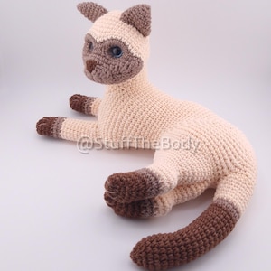 Può includere: Un peluche a forma di gatto siamese all'uncinetto, nei colori crema, marrone e beige. Il gatto è sdraiato con le zampe distese. Ha gli occhi blu e segni marroni sul viso, sulle orecchie, sulle zampe e sulla coda.