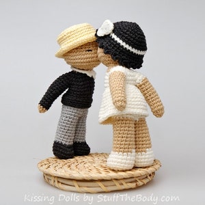 Peut inclure: Deux poup&eacute;es en crochet s'embrassant. La poup&eacute;e de gauche porte un pull noir, un pantalon gris et un chapeau jaune. La poup&eacute;e de droite porte une robe blanche et un chapeau noir avec un ruban blanc. Les poup&eacute;es sont debout sur une base en osier.