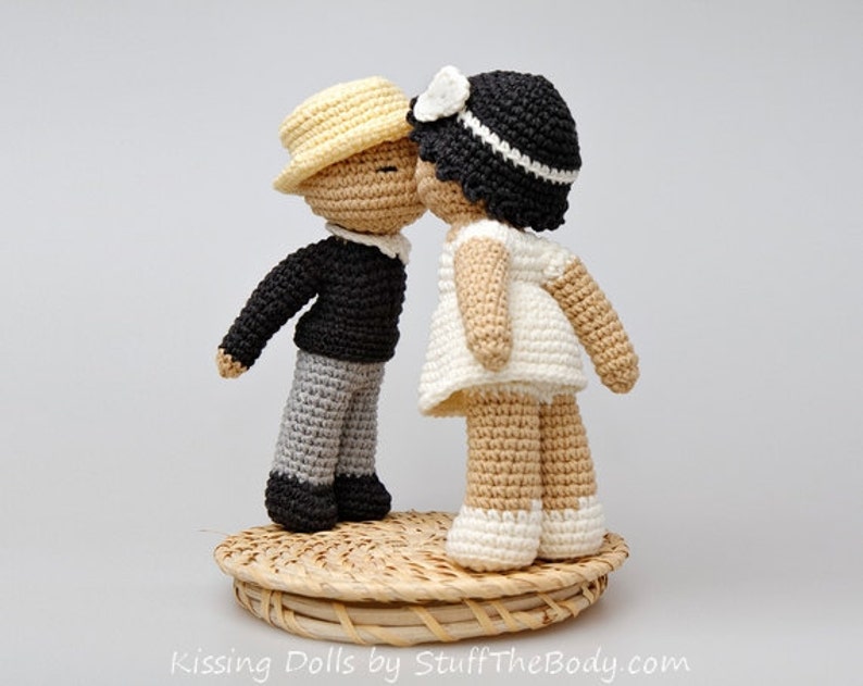 Peut inclure: Deux poup&eacute;es en crochet s'embrassant. La poup&eacute;e de gauche porte un pull noir, un pantalon gris et un chapeau jaune. La poup&eacute;e de droite porte une robe blanche et un chapeau noir avec un ruban blanc. Les poup&eacute;es sont debout sur une base en osier.