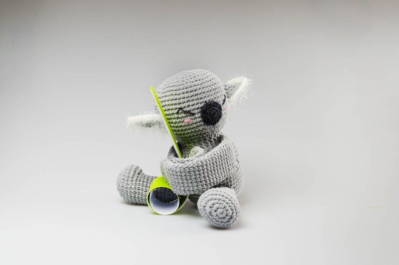 May include: A gray crocheted koala plush toy holding a green and white striped tube. The koala has black eyes and pink cheeks.