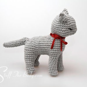May include: A gray crocheted cat toy with a red ribbon around its neck. The cat is standing on a white surface.