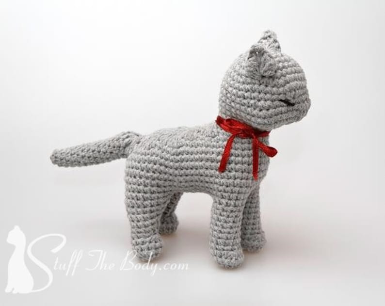 May include: A gray crocheted cat toy with a red ribbon around its neck. The cat is standing on a white surface.
