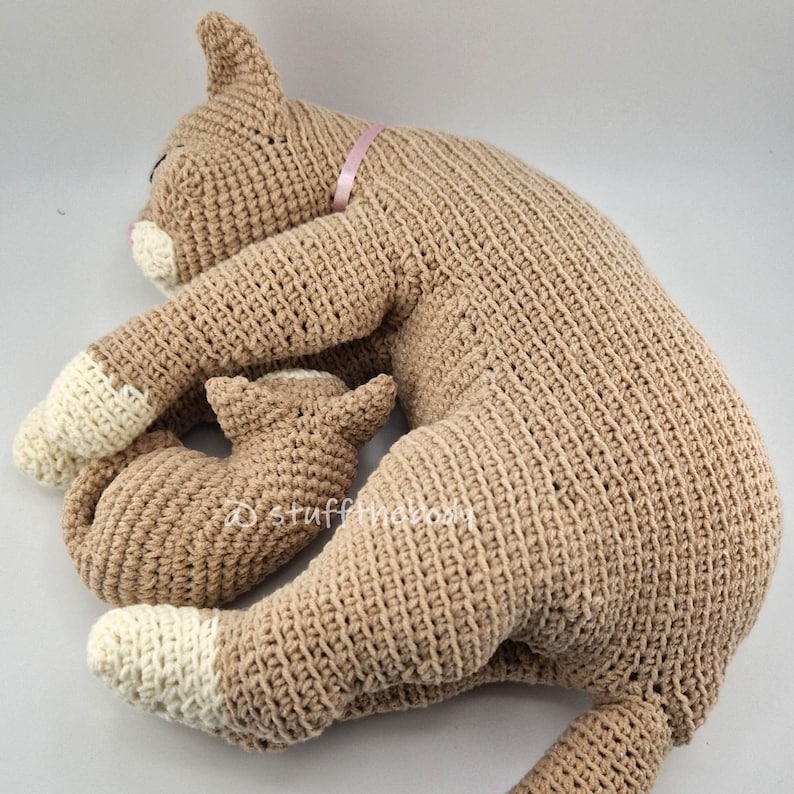 May include: A large, crocheted cat plush in a light brown color, with a smaller matching kitten. The cat has white paws and a pink ribbon around its neck. The kitten is nestled next to the larger cat.
