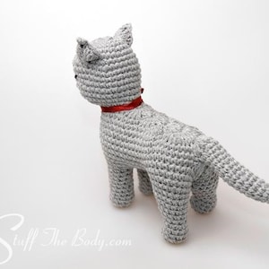 May include: A gray crocheted cat toy with a red ribbon around its neck. The cat is standing on a white surface.