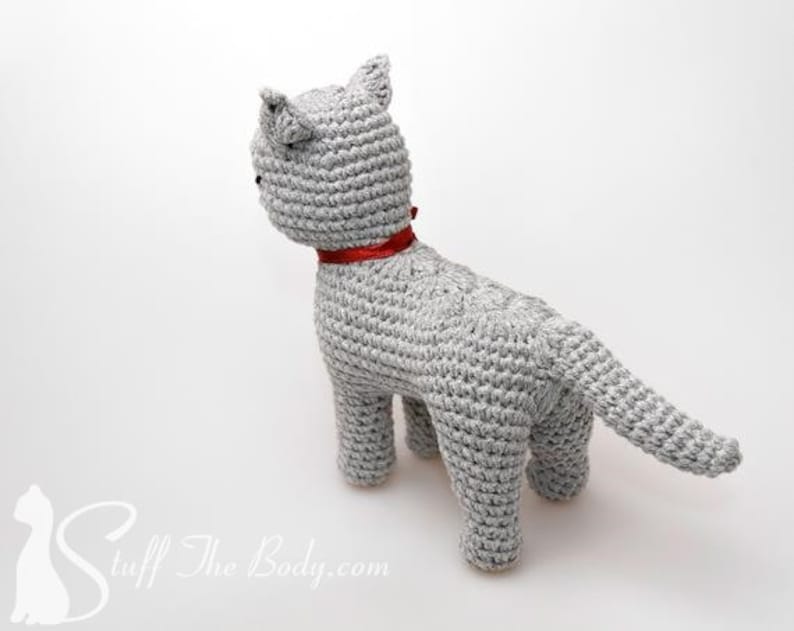 May include: A gray crocheted cat toy with a red ribbon around its neck. The cat is standing on a white surface.