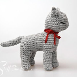 May include: A gray crocheted cat toy with a red bow around its neck. The cat is standing on a white surface.