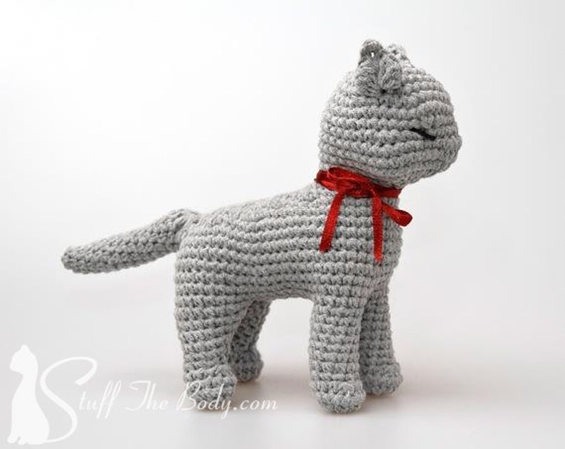 May include: A gray crocheted cat toy with a red bow around its neck. The cat is standing on a white surface.