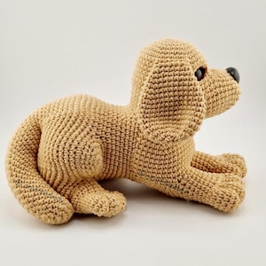 May include: A crocheted golden retriever puppy toy in a lying down position. The toy is made with beige yarn and has a brown nose and black eyes.
