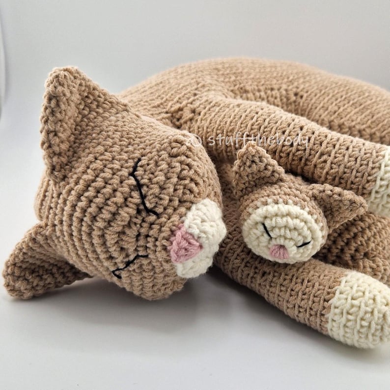 May include: A crocheted cat plush toy in shades of tan and cream. The cat is lying down, with its eyes closed and a pink nose. The plush has a soft, textured appearance, and the details are carefully stitched.