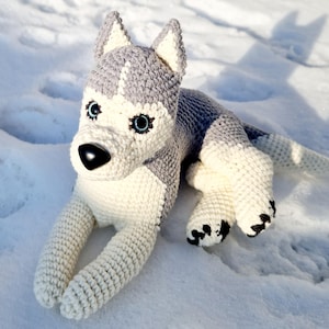 May include: A crocheted husky dog plush toy with white and gray fur, blue eyes, and black nose. The toy is lying on a white snowy surface.