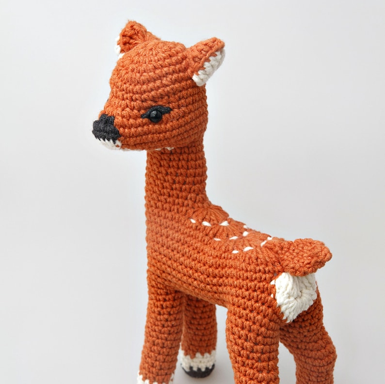 A crocheted amigurumi fawn with orange-brown body, white spots on its back, white accents on ears, hooves, and tail, and a black nose