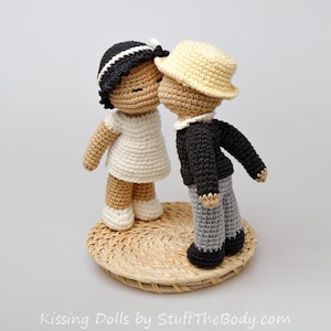 Peut inclure: Deux poup&eacute;es en crochet s'embrassant. La poup&eacute;e de gauche porte une robe blanche et a les cheveux noirs. La poup&eacute;e de droite porte une veste noire, un pantalon gris et un chapeau jaune. Les poup&eacute;es sont debout sur une base tiss&eacute;e.
