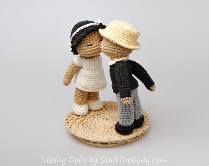 Peut inclure: Deux poup&eacute;es en crochet s'embrassant. La poup&eacute;e de gauche porte une robe blanche et a les cheveux noirs. La poup&eacute;e de droite porte une veste noire, un pantalon gris et un chapeau jaune. Les poup&eacute;es sont debout sur une base tiss&eacute;e.