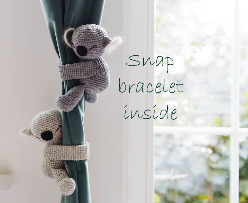 Two crocheted amigurumi koalas with snap bracelets inside their arms, wrapped around a curtain as cute curtain tiebacks