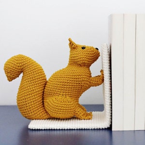 Könnte beinhalten: Ein gehäkelter Eichhörnchen-Buchstütze in senfgelber Farbe, die einen Stapel weißer Bücher hält.