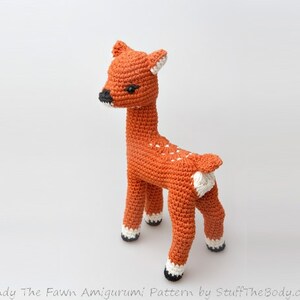 May include: A crocheted fawn amigurumi toy with brown fur, white spots, and black hooves. The fawn is standing on a white background.