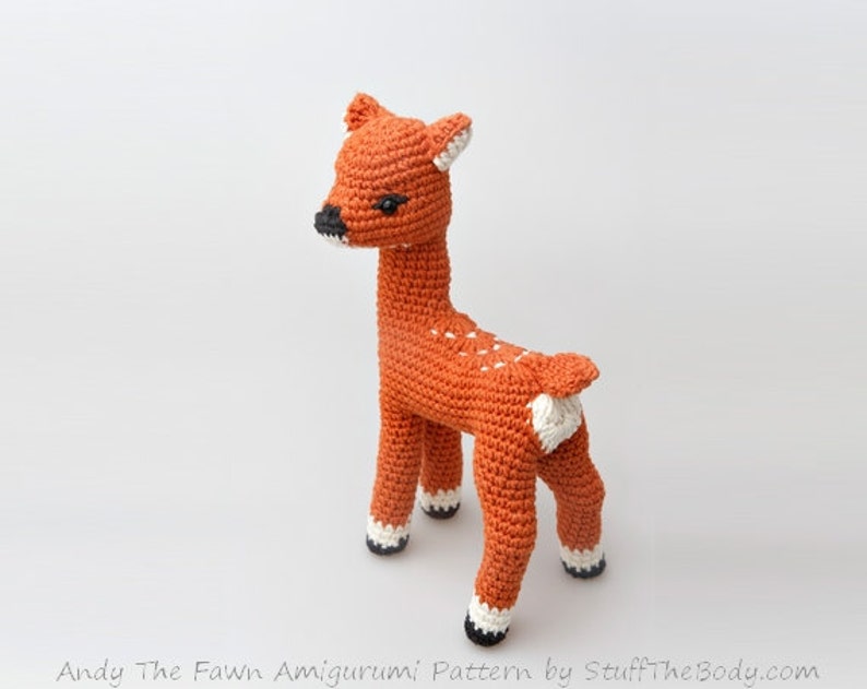 May include: A crocheted fawn amigurumi toy with brown fur, white spots, and black hooves. The fawn is standing on a white background.