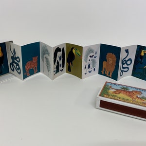 May include: A matchbox with a colorful illustration of a tiger, palm trees, and the text "Palm & Tiger" and "Melaka Match Factory". The matchbox is open, revealing a small booklet with illustrations of jungle animals, including a toucan, snake, tiger, palm leaves, beetles, and a lion.