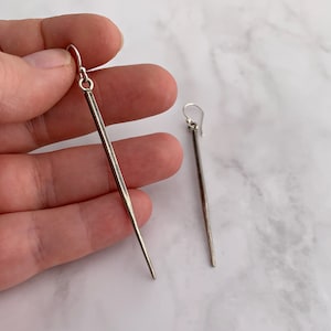 May include: A pair of long, silver earrings with a simple, straight design. The earrings are made of a textured metal and have a slightly pointed tip.