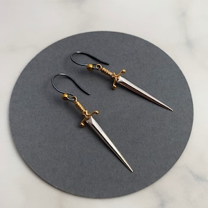 May include: A pair of silver dagger earrings with gold accents and black wire hooks. The daggers are detailed with a twisted rope design.