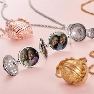 Puede incluir: Varios relicarios con fotos sobre una superficie rosa. Los relicarios están abiertos, mostrando fotos de personas. Los relicarios son plateados y dorados, con diseños intrincados. Los collares tienen cadenas plateadas y doradas.