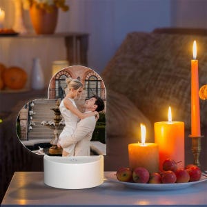 May include: A heart-shaped acrylic plaque featuring a printed photograph of a couple embracing in front of a stone fountain. The plaque is mounted on a circular white base with a power button, illuminated by two nearby orange pillar candles.