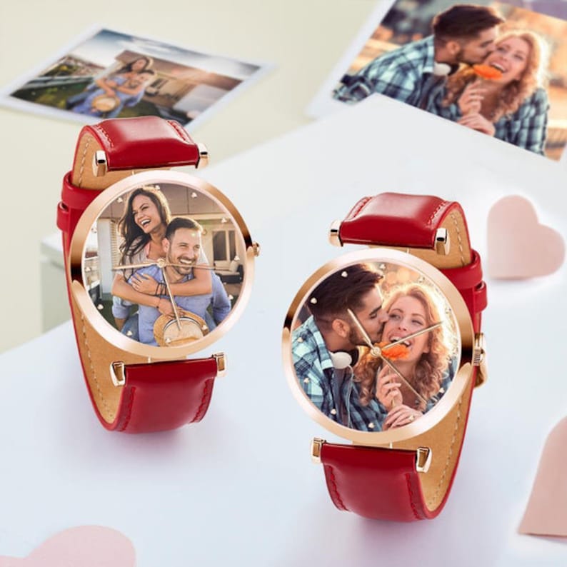 May include: Two red leather strap watches with gold-tone cases. The watch faces display personalized photos of couples. The watches are on a white surface with heart-shaped decorations and photos in the background.
