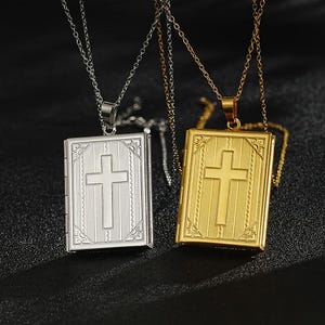 Puede incluir: Dos collares con colgante con un diseño de medallón en forma de libro. Uno es plateado, el otro dorado. Cada medallón presenta una cruz en la parte delantera y cuelga de una delicada cadena. El fondo es oscuro.