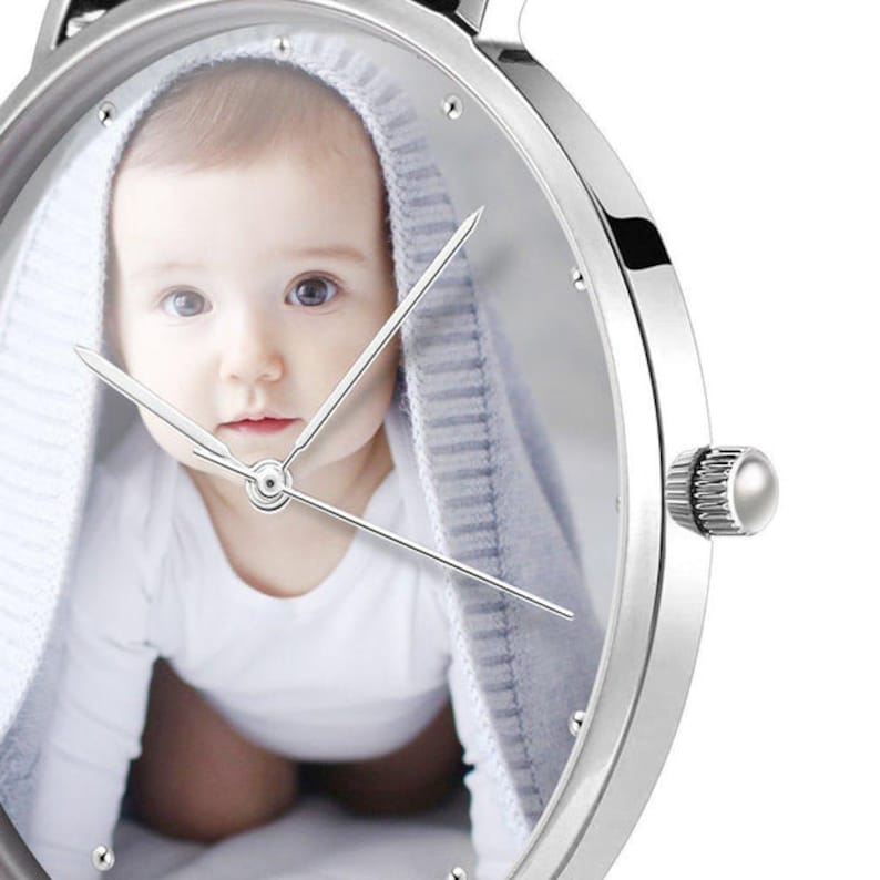 May include: A silver wristwatch with a clear face featuring a baby wrapped in a light blue blanket. The watch has silver hands and a silver bezel. The watch is a unique and sentimental accessory.