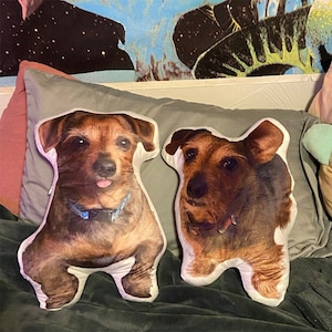 May include: Two brown and white dog-shaped pillows with a white background. The pillows are on a gray and brown surface.